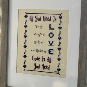 May include: A framed cross-stitch design featuring the text "All You Need Is Love" with mathematical equations forming the letters "LOVE". The design is on a beige background with purple and black thread.
