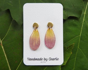 May include: A pair of handmade earrings featuring gold studs and teardrop pendants. The pendants display a yellow to pink gradient. The earrings are presented on a white card with the text "Handmade by Charlie EST. 2014".