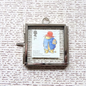 Op de afbeelding: Een zilveren ingelijst plaatje van een Paddington Bear-postzegel. De postzegel is rood, wit en blauw en toont Paddington Bear met een rode hoed en een pot marmelade in zijn hand. De tekst op de postzegel luidt "More About Paddington by Michael Bond Illustrated by Peggy Fortnum".