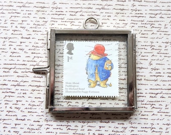 May include: A silver framed picture of a Paddington Bear stamp. The stamp is red, white, and blue and features Paddington Bear wearing a red hat and holding a jar of marmalade. The text on the stamp reads "More About Paddington by Michael Bond Illustrated by Peggy Fortnum".
