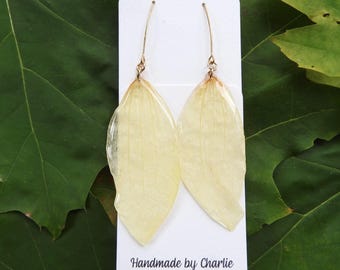 May include: A pair of drop earrings featuring pale yellow, translucent leaf-shaped pendants suspended from thin gold-toned metal hooks. The earrings are attached to a white rectangular display card with the text Handmade by Charlie est. 2014.