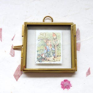 Op de afbeelding: Een gouden metalen frame met een glazen voorkant toont een vintage Japanse postzegel met een konijn dat een wortel eet met een roodborstje dat op een tuingereedschap zit. De postzegel heeft de tekst "80 NIPPON".