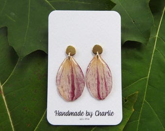 May include: A pair of drop earrings featuring teardrop-shaped petals with cream and magenta veining, attached to small gold-tone circular studs. The earrings are displayed on a white rectangular card with the text Handmade by Charlie EST. 2014.
