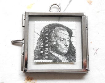 May include: A square silver-coloured metal frame with a glass front and back, containing a postage stamp featuring a black and white portrait of Rembrandt. The stamp includes the text "NEDERLAND", "0,39 euro", and "REMBRANDT". A small metal loop is attached to the top.