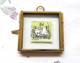May include: A square brass frame with a hinged side and a top loop, containing a light green Japanese postage stamp. The stamp features a white cartoon character lying in a field of colourful flowers with the text NIPPON 84 printed above.