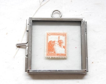 May include: A square silver-toned metal frame with clear glass panels containing a vintage orange postage stamp. The stamp features the number 5, the word PALESTINE, and an illustration of a stone fortress. A small metal ring is attached to the top.