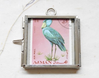 May include: A square silver-toned metal frame containing a postage stamp featuring a blue shoebill bird on a pink background. The stamp includes the text 1 RI, AIR MAIL, BALAENICEPS REX, and AJMAN in English and Arabic script with a circular postmark.