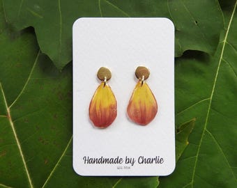 May include: Handmade teardrop earrings with a yellow to red gradient, and small, round, gold-coloured studs. The white card displays "Handmade by Charlie EST. 2014". The earrings are presented against a green leaf background.