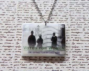 May include: A rectangular pendant necklace featuring a black and white historical image of a suffragette holding a sign that reads Give Women The Vote This Session. The text Votes For Women and Lone Suffragette In Whitehall 1908 is printed below.
