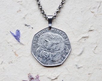 May include: A silver-coloured heptagonal coin pendant featuring an embossed illustration of Mrs. Tiggy-Winkle, a hedgehog character, with the text MRS. TIGGY-WINKLE inscribed along the top edge, suspended from a matching silver-coloured metal link chain.