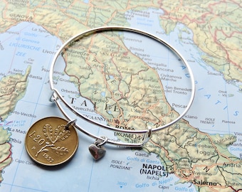 May include: A silver-toned expandable wire bangle bracelet featuring a circular bronze-coloured coin charm marked with the text L.20 and 1957, alongside a small silver-toned heart-shaped charm, resting on a detailed map of Italy.