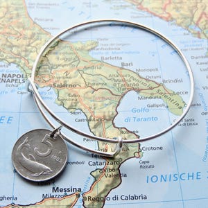 May include: A silver-toned expandable wire bangle bracelet featuring a circular metal charm. The charm is a coin displaying the number 5 and a dolphin illustration, with the year 1953 visible on its surface. The bracelet is circular in shape.