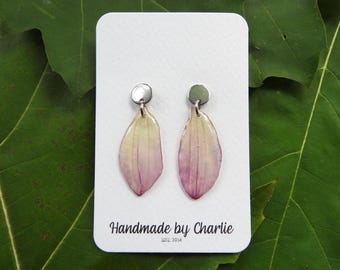May include: A pair of drop earrings featuring translucent, teardrop-shaped petals with a gradient from pale yellow at the top to soft pink at the bottom. Each petal hangs from a small, circular silver-toned stud. The text reads Handmade by Charlie EST. 2014.
