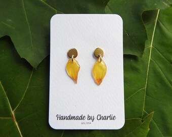 May include: Handmade earrings with yellow petal-shaped pendants and brass fittings. The earrings are presented on a white card with the text "Handmade by Charlie EST. 2014". The background is a green leaf.