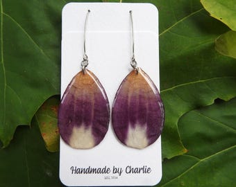 May include: A pair of teardrop-shaped earrings featuring preserved flower petals with a gradient of deep purple, tan, and white. The earrings hang from silver-toned metal hooks and are displayed on a white card labelled Handmade by Charlie EST. 2014.
