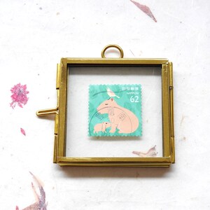 Op de afbeelding: Een gouden frame met een glazen voorkant toont een vintage Japanse postzegel met een capybara met twee jonge capybara's en een kleine vogel op een groen blad. De postzegel is gelabeld "NIPPON 62".