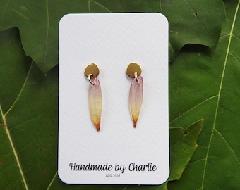 May include: Handmade earrings with gold studs and petal-shaped dangles in pink, yellow, and brown. The earrings are displayed on a white card with the text "Handmade by Charlie" and the year "EST. 2014". The background is a green leaf.