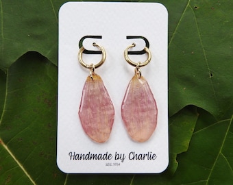 May include: A pair of gold hoop earrings featuring dangling, translucent, pink and yellow pressed flower petals. The earrings are attached to a white rectangular display card with the text Handmade by Charlie EST. 2014 printed in black.