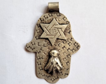 Pode incluir: Amuleto de prata em forma de mão de Hamsa com uma estrela de seis pontas no centro. O amuleto tem uma superfície texturizada com um pequeno elemento decorativo na parte inferior.