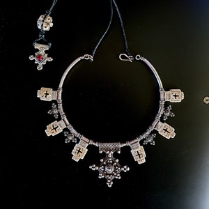 Peut inclure: Un collier en argent avec un grand pendentif en forme de croix et de plus petits charmes en forme de croix. Le collier est fait d'un fil d'argent et a un cordon noir attaché.