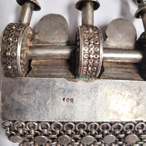 May include: Close-up of a silver necklace with a detailed design. The necklace has a chain with small round links and a pendant with a decorative design. The pendant has a silver bar with the inscription "W 08".
