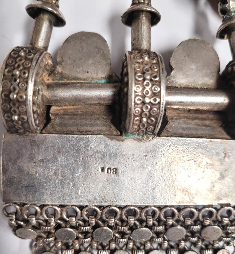 May include: Close-up of a silver necklace with a detailed design. The necklace has a chain with small round links and a pendant with a decorative design. The pendant has a silver bar with the inscription "W 08".