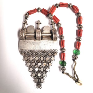 May include: A silver pendant necklace with a triangular design and a silver chain. The pendant features a detailed design with a small silver charm. The necklace is strung with red coral beads and green glass beads.