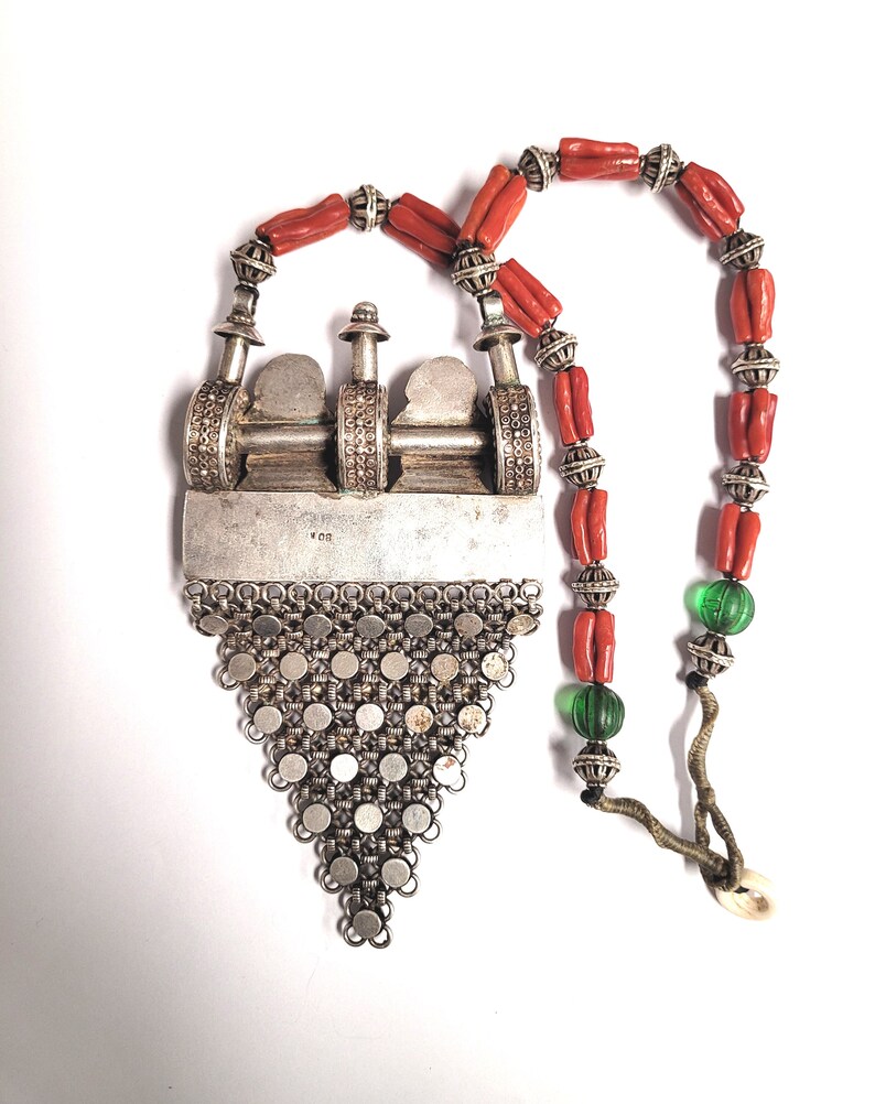 May include: A silver pendant necklace with a triangular design and a silver chain. The pendant features a detailed design with a small silver charm. The necklace is strung with red coral beads and green glass beads.