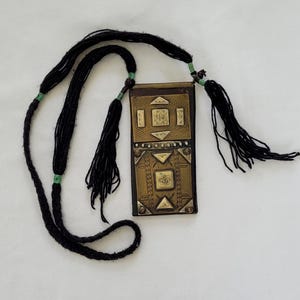 May include: A rectangular pendant with a decorative brass front and a black cord. The brass is adorned with geometric patterns and shapes. The pendant is attached to a long black cord with tassels and green accents.