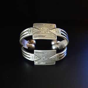 May include: A silver bracelet with intricate geometric designs. The bracelet is made of three bands with a decorative square piece on each end.