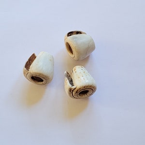 Può includere: Tre perline di ceramica bianca con un foro al centro. Le perline hanno una superficie ruvida e testurizzata e sono leggermente irregolari.