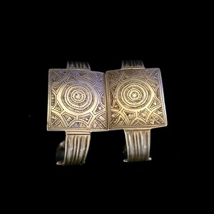 May include: Two silver-toned cuff bracelets with rectangular centerpieces. Each centerpiece features an intricate geometric pattern with a central circular design. The bracelets have a vintage or tribal aesthetic, set against a black background.