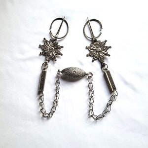 May include: A pair of silver-toned metal brooches with ornate floral designs and a connecting chain with a decorative oval pendant.