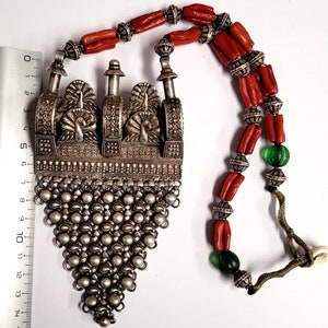 May include: A silver necklace with a large pendant featuring three peacocks. The pendant is made of intricate silverwork and has a chain of small silver beads. The necklace is strung with red coral beads and green glass beads.