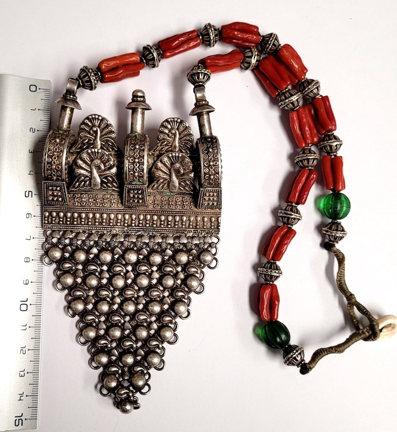 May include: A silver necklace with a large pendant featuring three peacocks. The pendant is made of intricate silverwork and has a chain of small silver beads. The necklace is strung with red coral beads and green glass beads.