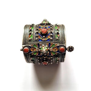 May include: A silver cuff bracelet with intricate detailing and colorful enamel accents. The bracelet features red, green, blue, and yellow enamel, with small, round embellishments and a decorative clasp. The design is ornate and eye-catching.