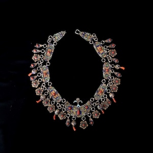 May include: A silver necklace with intricate enamel work and coral accents. The necklace features a central pendant with a crescent moon design and multiple dangling charms.