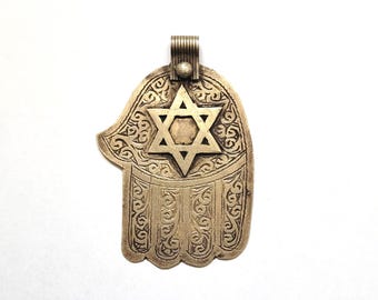 Pode incluir: Pingente de mão Hamsa prateado com um recorte da Estrela de David. O pingente apresenta intrincados trabalhos de rolagem e um laço para pendurar. O design é simétrico e detalhado, com uma estética vintage.