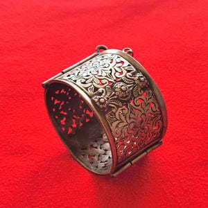 May include: Silver cuff bracelet with an ornate floral cut-out design. The bracelet displays detailed patterns of flowers and leaves, with a hinged clasp. The background is a solid red colour.