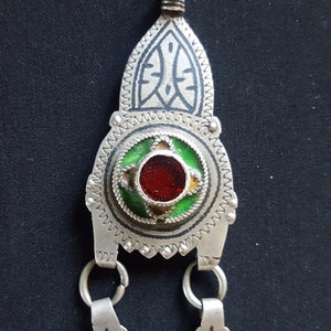 以下が含まれることがあります： 緑と金のエナメル模様と中央に赤いガラスの石がついた銀製のペンダント。ペンダントには、幾何学模様のチャームが2つぶら下がっている。