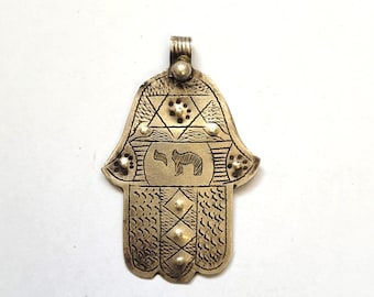 Pode incluir: Pingente de mão Hamsa prateado com detalhes intrincados gravados. O pingente apresenta uma Estrela de Davi, um símbolo e padrões geométricos. O amuleto em forma de mão é projetado para joias e adorno espiritual.