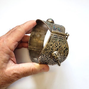 May include: A silver bracelet with intricate floral designs and blue and green enamel accents. The bracelet has a hinged clasp and a decorative bell charm.