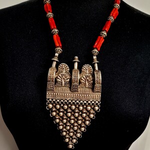 May include: A silver necklace with a large, ornate pendant featuring peacocks and a fringe of silver beads. The necklace is strung with red coral beads.