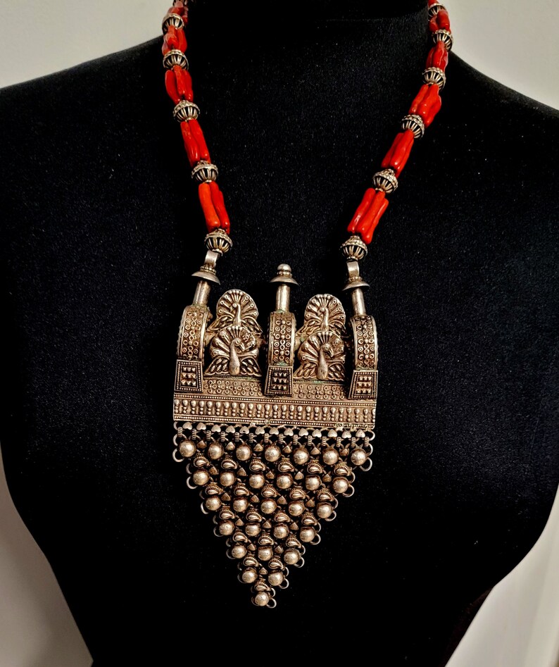 May include: A silver necklace with a large, ornate pendant featuring peacocks and a fringe of silver beads. The necklace is strung with red coral beads.
