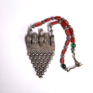 May include: A silver necklace with a large, ornate pendant featuring a geometric design and a chain-like fringe. The necklace is strung with red coral beads and green glass beads.