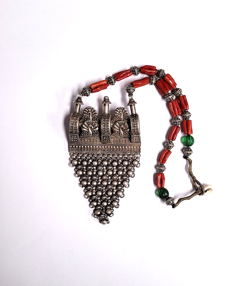May include: A silver necklace with a large, ornate pendant featuring a geometric design and a chain-like fringe. The necklace is strung with red coral beads and green glass beads.
