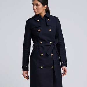 May include: A dark navy blue double-breasted trench coat featuring a classic collar, long sleeves with buckled cuffs, and a matching waist tie belt. The coat is adorned with two vertical rows of polished gold-tone buttons down the front.