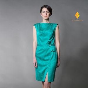Op de afbeelding: Een vrouw draagt een tealgroene, mouwloze jurk met een col en een split aan de zijkant. De jurk heeft een subtiel patroon en een riem in de taille.