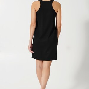 May include: A black sleeveless dress with a scoop neck and a straight silhouette. The dress is worn by a person with long dark hair, walking away from the camera.