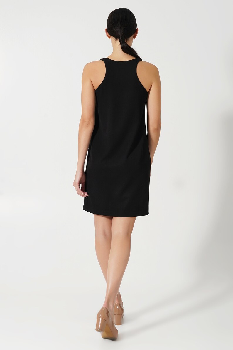 May include: A black sleeveless dress with a scoop neck and a straight silhouette. The dress is worn by a person with long dark hair, walking away from the camera.